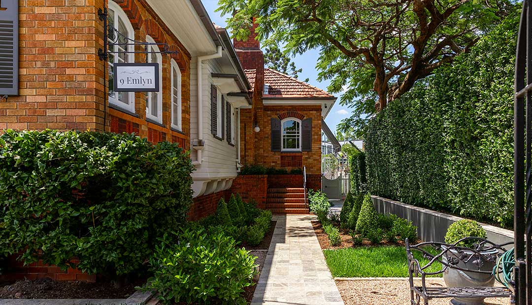 A brick house with a "9 Emlyn" sign, lush greenery, and a tidy walkway on Emlyn Street, Brisbane.